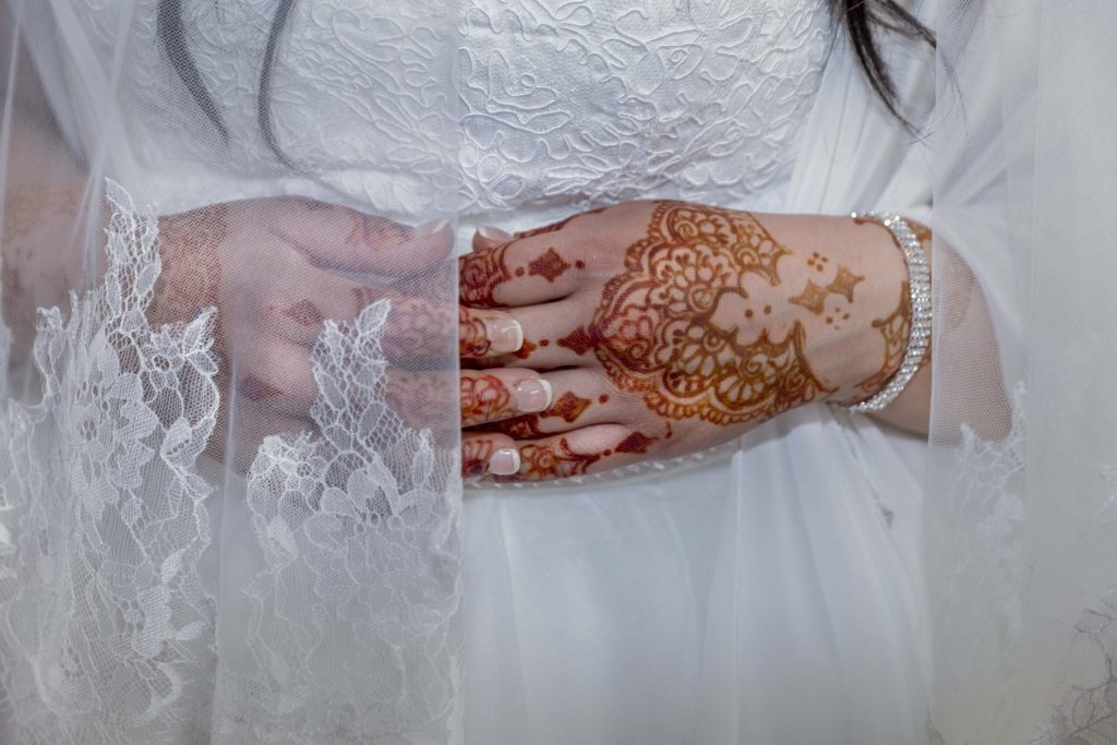 Close up shot of a bride in a white lace wedding gown and lace veil. Her hands, intricately decorated in henna, are folded over her midsection.